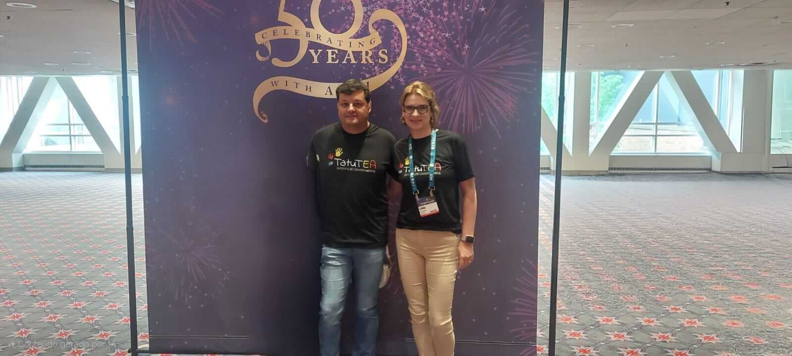 Lívia e Fernando com camisetas do TatuTEA no painel de 50 anos da ABAI, em Filadélfia